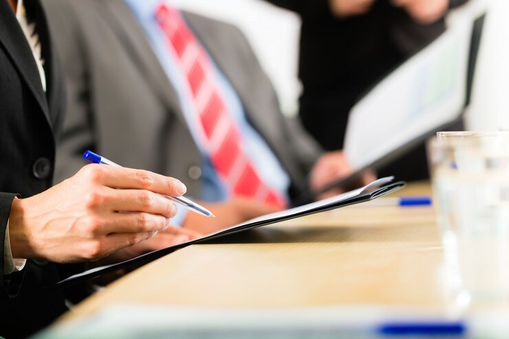 A close-up of a person in a suit writing on a clipboard during a formal meeting